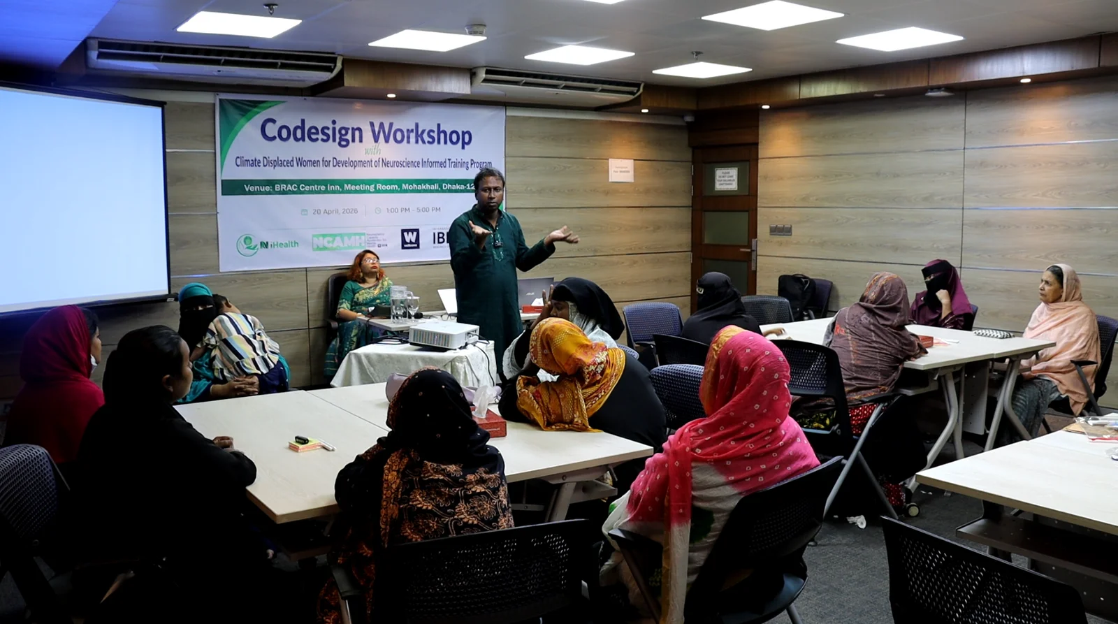 Women seated at tables facing a facilitator, beneath a banner reading Codesign Workshop with Climate Displaced Women for Development of Neuroscience Informed Training Program